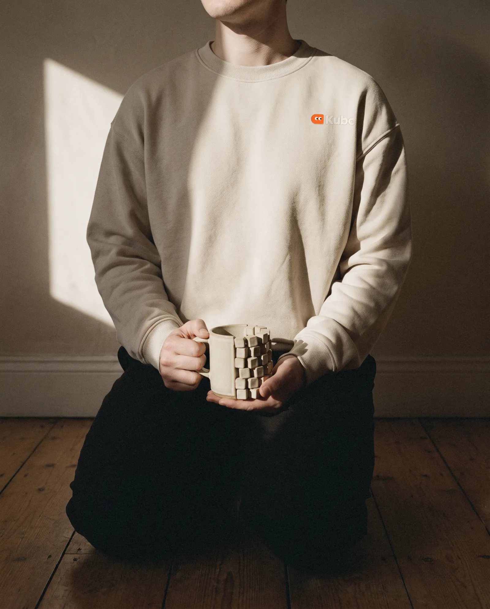 An oat crewneck with the orange mark embroidered on the chest, held with a half-cube mug