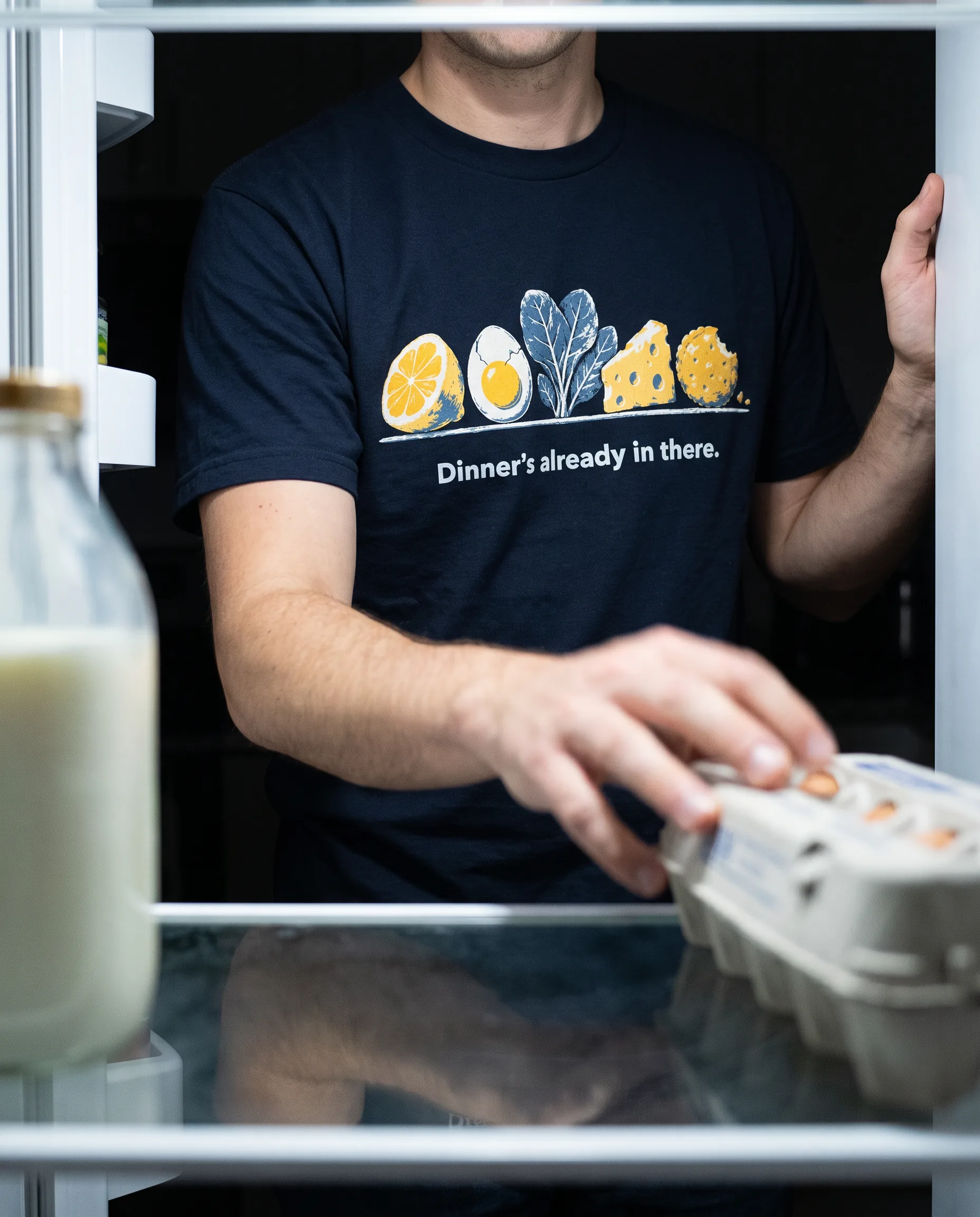 A person in a navy Leftly tee at an open fridge, the print a row of lemon, egg, spinach, cheese and a bitten biscuit