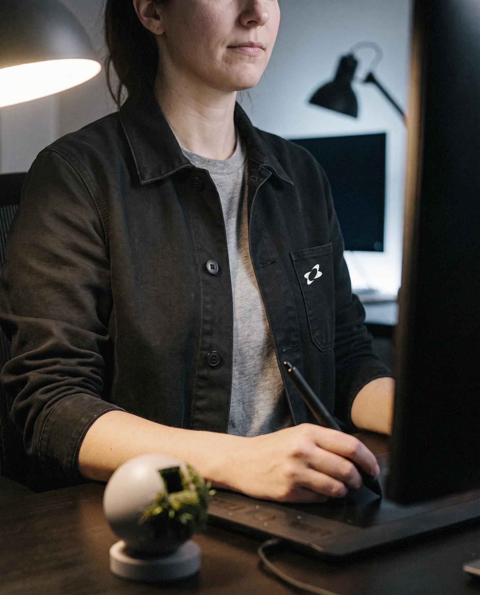 A game artist at her desk in a black chore jacket with the Texel mark on the pocket, a moss shader ball beside her